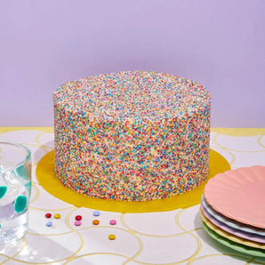 Pinata Cake covered in colorful rainbow sprinkles on yellow board, ready to delight with mini Smarties inside.